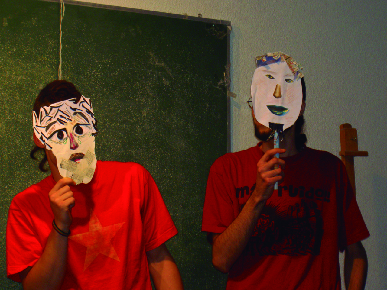 Dos personas con camisetas rojas ocultan sus rostros tras dos caretas confeccionadas manualmente en un ámbito de aula de teatro.